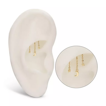 A gold-colored stud earring with chain links leading to a crescent moon and stars dangle, on display in the hidden conch piercing of a model ear.